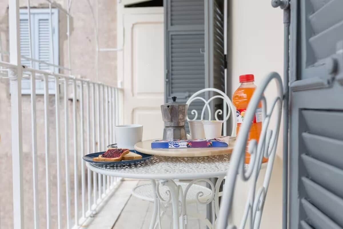 A balcony with a white metal table, a coffee pot, a plate with food, a cup, and a bottle of orange juice.