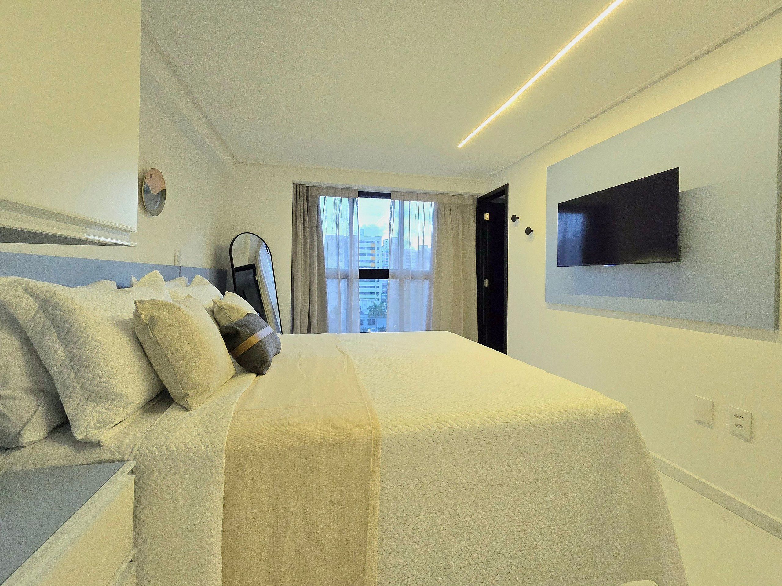 A hotel room with a bed, a flat-screen TV mounted on the wall, and a window with curtains.