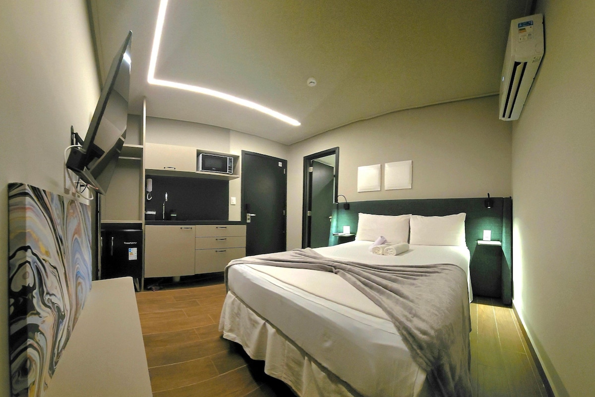 Habitación de hotel con cama, mesitas de noche, escritorio con silla, microondas, refrigerador y televisor de pantalla plana montado en la pared.