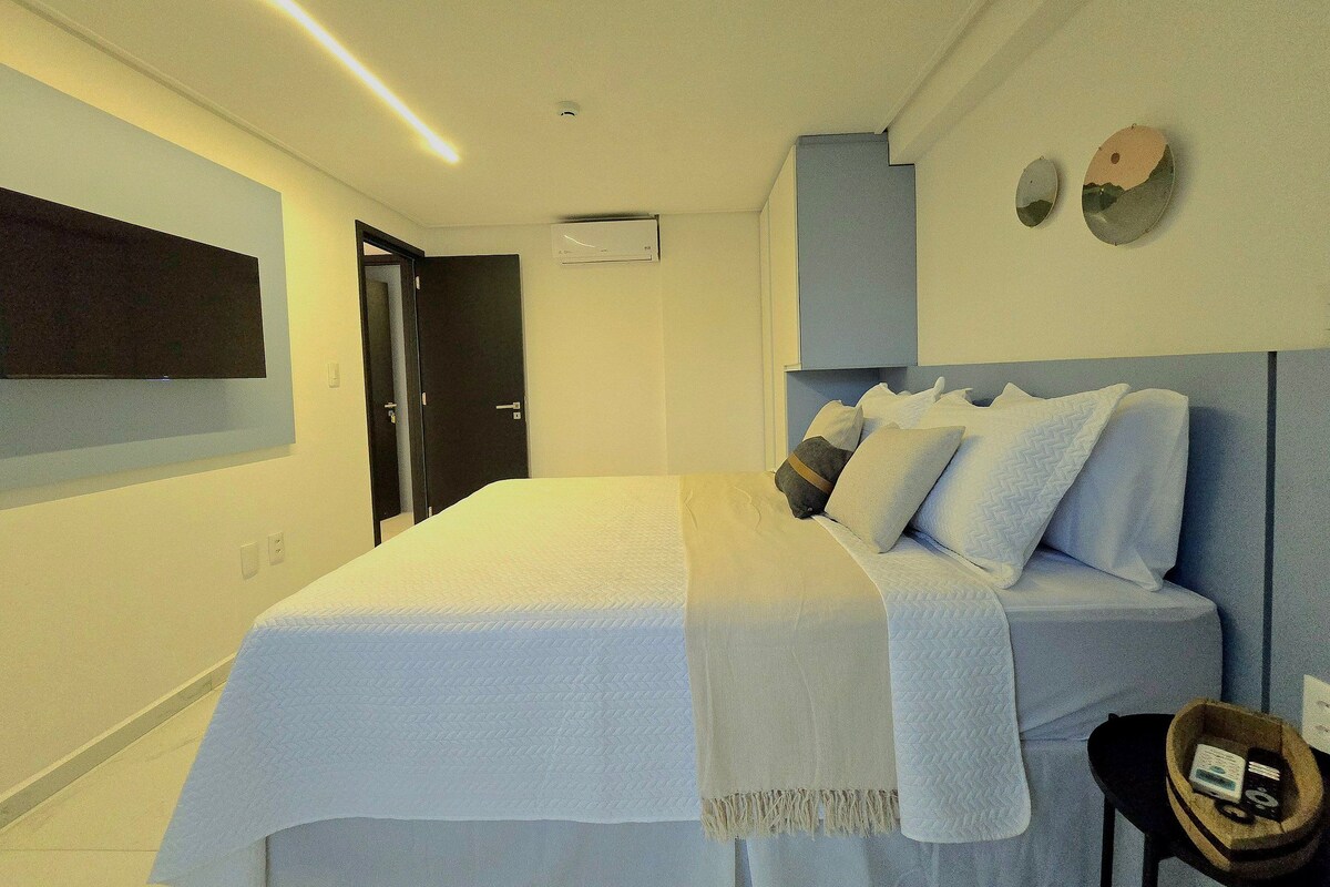 Un dormitorio moderno con una cama grande, una mesita de noche, un televisor montado en la pared y una puerta.