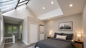 Une chambre avec un grand lit, deux tables de chevet avec des lampes, un ventilateur de plafond, un tableau représentant un paysage urbain encadré et un puits de lumière.