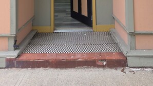 Une entrée d’immeuble avec un sol à carreaux, une porte en arc de cercle et, au fond, une cuisine visible.