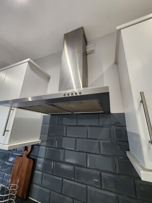 A kitchen with a stainless steel range hood, white cabinets, and a black tiled backsplash. - The Walnut (South Ockendon)