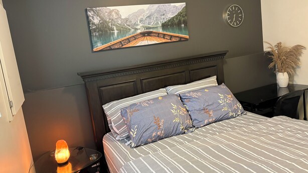 A bedroom with a bed, a nightstand, a lamp, a wall clock, and a painting. - Two bedroom cozy apartment in central location, 15 minute from historic downtown (St. John's)