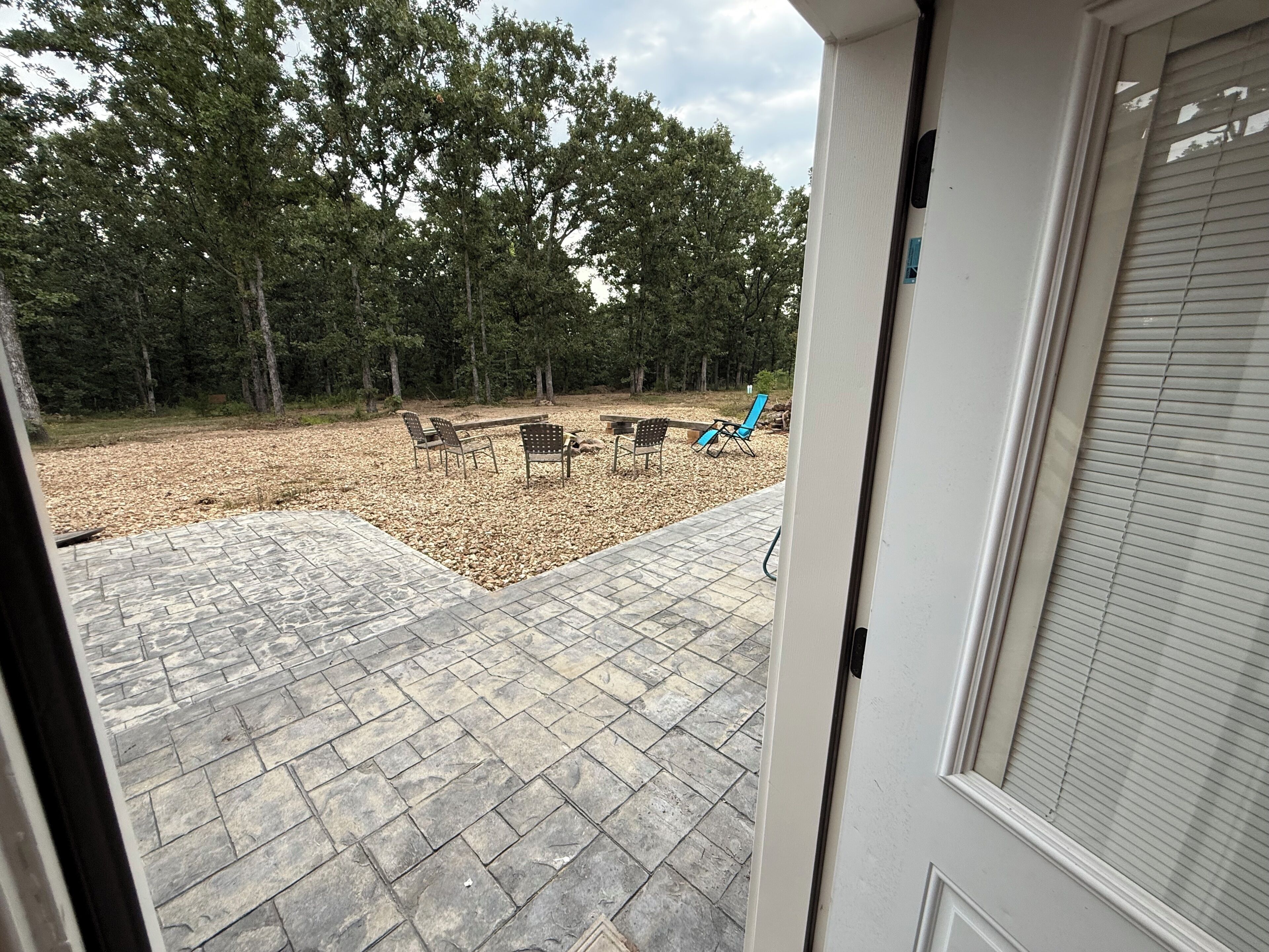 An outdoor patio with stone flooring, a seating area with chairs, and a view of a forested area.