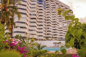 Een meerlaags hotel met balkons, een zwembad en weelderige begroeiing.