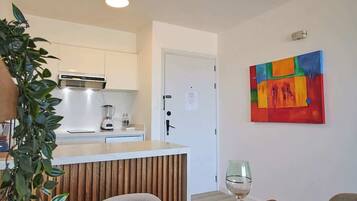 Een eethoek met een houten tafel voor vier personen, een wit kastje met keukenapparatuur, en een kleurrijk abstract schilderij aan de muur.