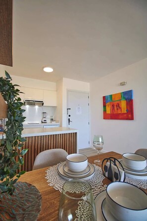 A dining area with a wooden table set for four, a white cabinet with kitchen appliances, and a colorful abstract painting on the wall. - Refugio en el Mar Despierta frente al Caribe (Porlamar)