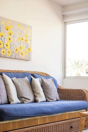 Un sofá de mimbre con cojines azules y varias almohadas, ubicado en una habitación con un ventanal grande y un cuadro floral colgado en la pared.