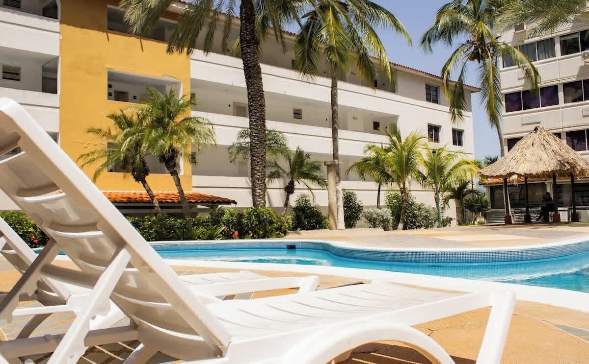 A swimming pool area with lounge chairs, palm trees, and a building with balconies.
