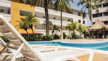 A swimming pool area with lounge chairs, palm trees, and a building with balconies.