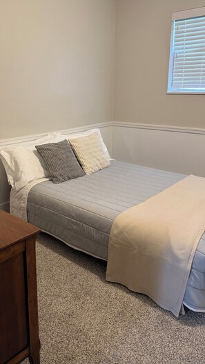 A bedroom with a bed, a window with blinds, and a wooden nightstand.