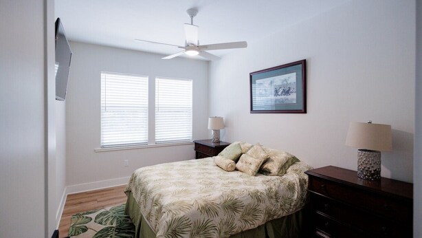 Quarto com cama, ventilador de teto, janela com persianas, mesa de cabeceira com abajur e um quadro pendurado na parede.