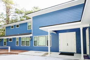 A two-story building with a blue exterior and white trim. There is a white door and a small porch area with a bench. - The Endzone | 6BR Near UF & Shands Sleeps 14 (Gainesville)