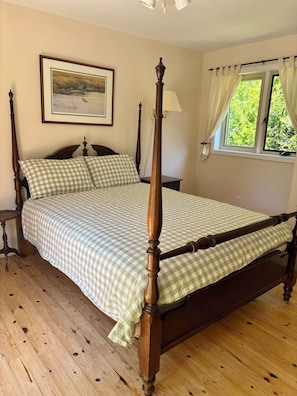 A four-poster bed with a checkered bedspread, a nightstand with a lamp, a framed picture on the wall, and a window with curtains.