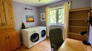 A laundry room with a washing machine, a chair, wooden cabinets, and a window with curtains.