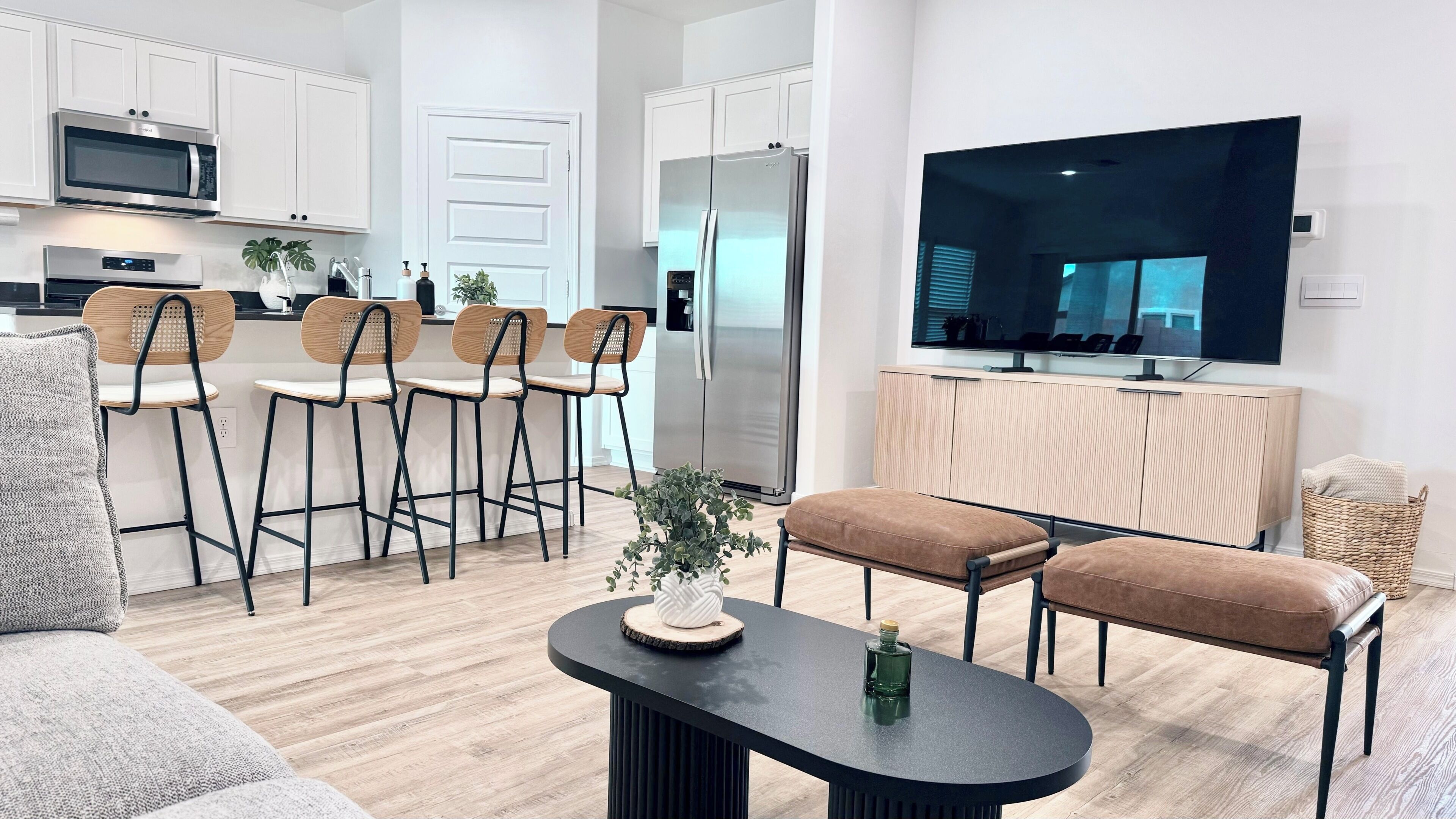 Une cuisine moderne avec des armoires blanches, un réfrigérateur en acier inoxydable et un four à micro-ondes. On y trouve un coin repas avec des chaises en bois et une table noire. Un grand téléviseur à écran plat est fixé au mur.