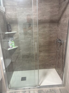 Eine gläserne Duschkabine mit Handbrause, Ablage für Toilettenartikel und sichtbarem Abfluss.
