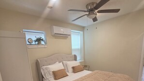 Quarto com cama, ventoinha no teto, ar condicionado, janela com persianas e relógio fixado na parede.