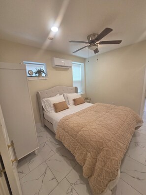 Ein Schlafzimmer mit einem Bett, einem Deckenventilator, einer Klimaanlage, einem Fenster mit Jalousien und einer Wanduhr.