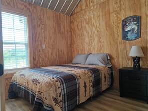 Une chambre avec un mur lambrissé en bois, un lit recouvert d’une couvre-lit à motifs, une table de chevet avec une lampe et un élément décoratif mural.