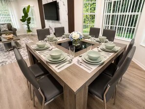 A dining area with a rectangular table set for six, surrounded by black chairs, with a large window and a plant in the background. - Family Getaway Near Disney • Pool & Hot Tub (Kissimmee)