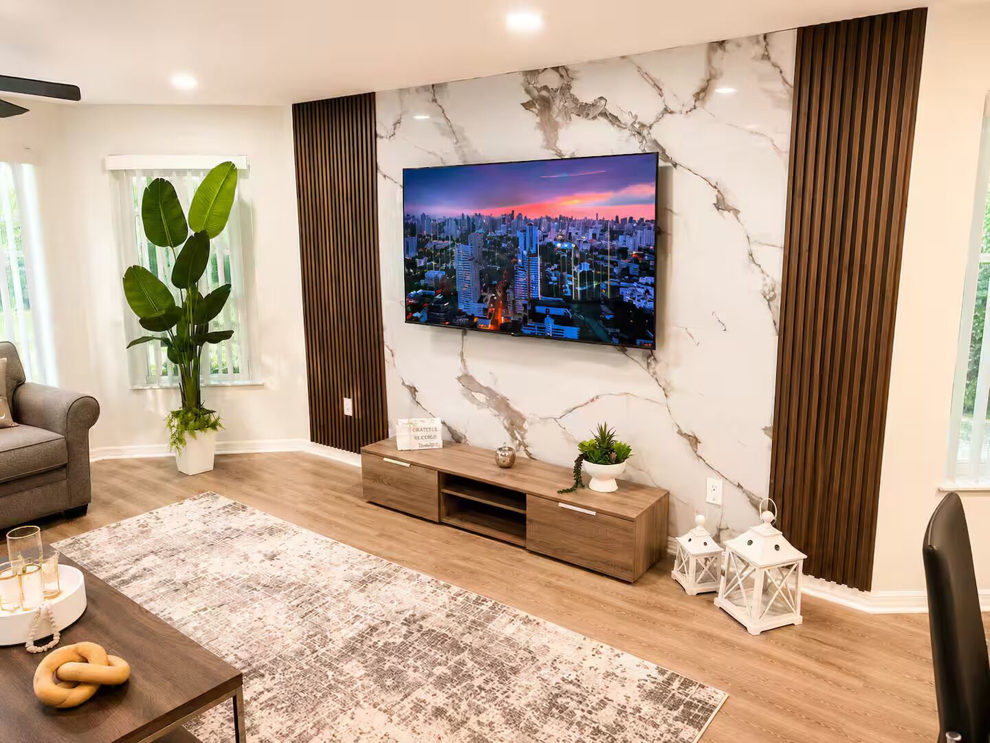 Un salon moderne doté d’un grand téléviseur à écran plat fixé sur un mur en marbre, d’un canapé gris, d’une table basse en bois et d’un tapis à motifs.