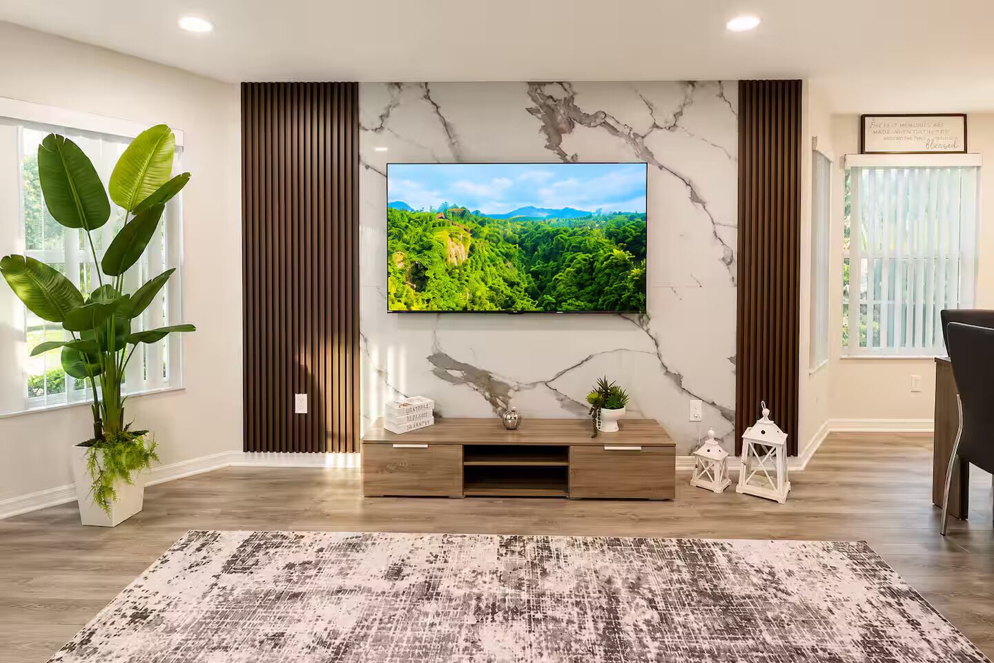 A modern living room with a flat-screen TV mounted on a marble wall, a wooden entertainment unit, a large potted plant, and a patterned rug.