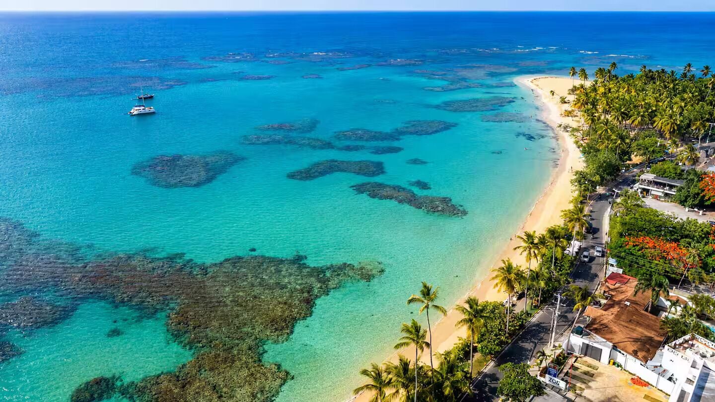 An aerial view of a coastal area with clear turquoise waters, a sandy beach, and a row of buildings with palm trees.