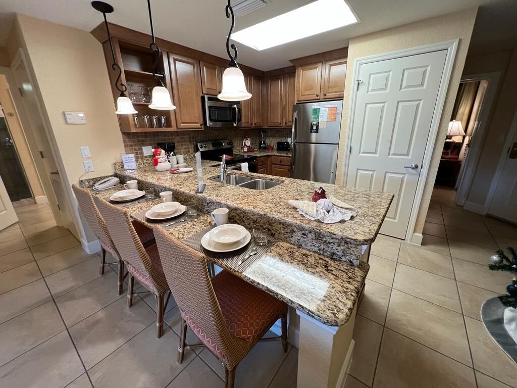 A kitchen with granite countertops, wooden cabinets, a stainless steel refrigerator, and a dining area with wicker chairs.