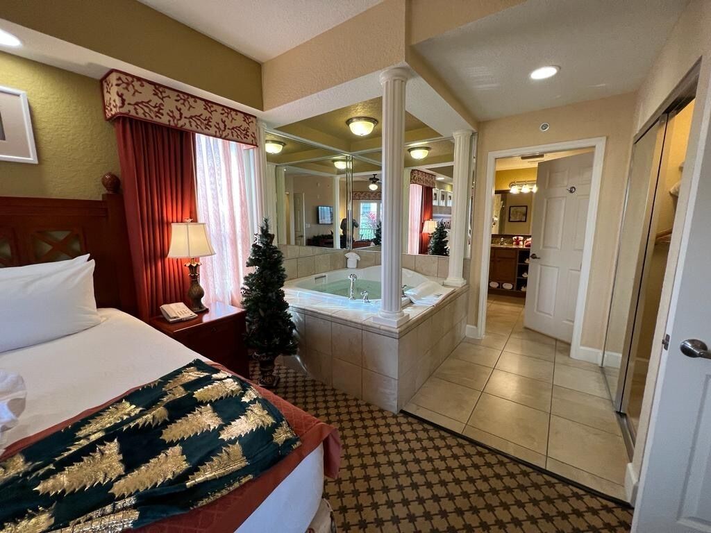 A hotel room with a bed, a bathroom with a bathtub, a mirror, and a door leading to another room.