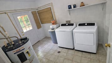 A laundry room with two white washing machines, a water heater, and a trash can.
