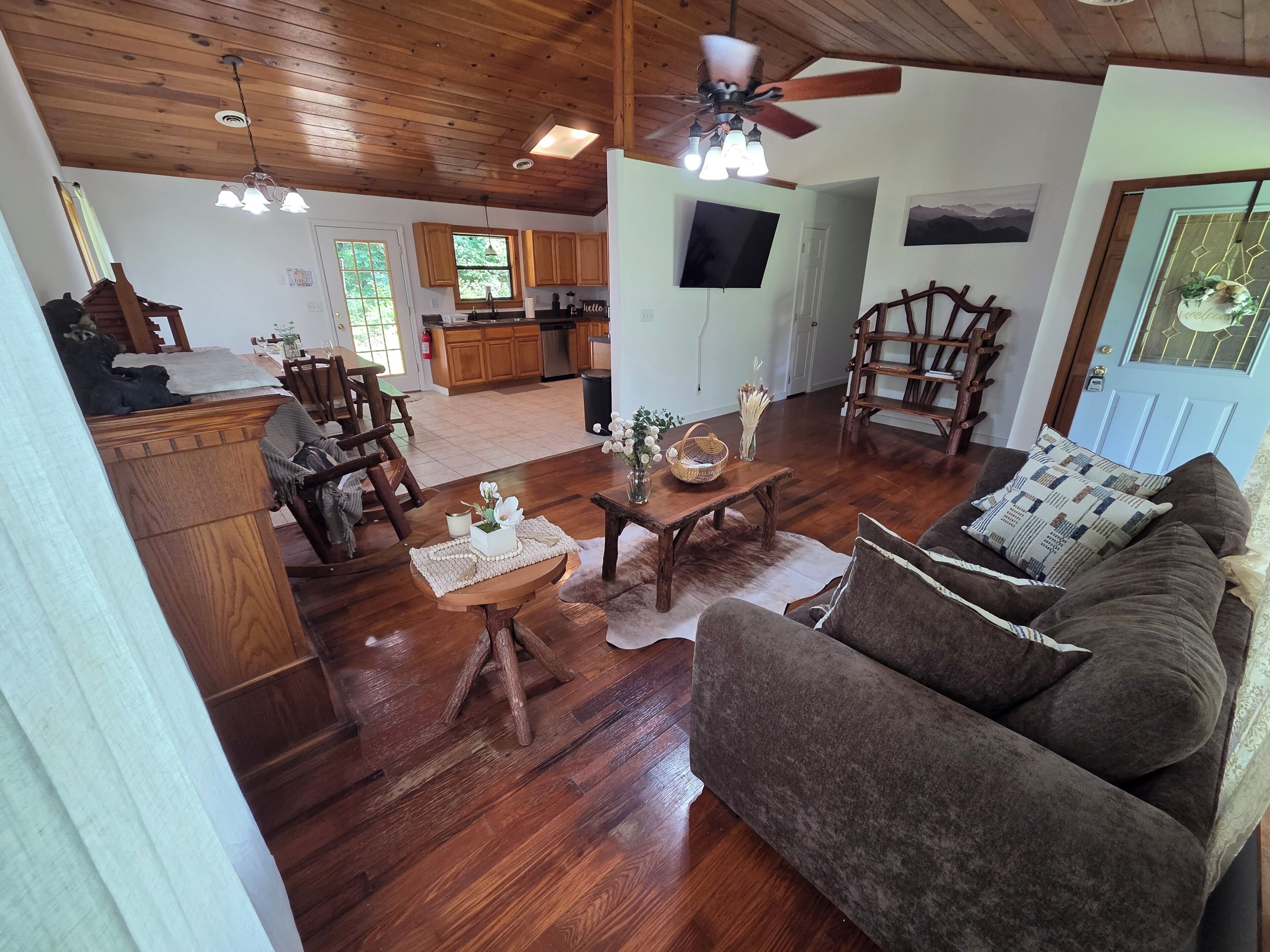 A spacious living room with a wooden ceiling, a large brown sofa, a coffee table, and a dining area with wooden chairs and a table.