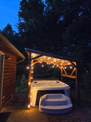 Un bain à remous sous une pergola en bois, illuminé de guirlandes lumineuses, entouré d’un chalet en bois.