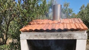Ein Backofen aus Ziegelsteinen im Freien mit Schornstein, der Essen zubereitet, und eine mit 'GRILL' beschriftete Holzkohlesack darunter.