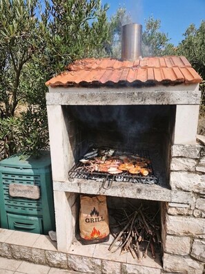 An outdoor brick oven with a chimney, cooking food, and a bag of charcoal labeled 'GRILL' underneath. - Apartment Duka "for 5" - 100m sandy beach + panoramic sea view + AirCon. & WiFi. (Lumbarda)