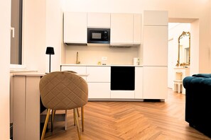 A modern kitchen with white cabinetry, a microwave, and a black oven. There is a chair with a quilted backrest and gold legs, and a dark blue sofa in the living area. - Dimora Smeraldo - an exclusive apartment just 10 minutes from the Amalfi Coast (Cava de' Tirreni)