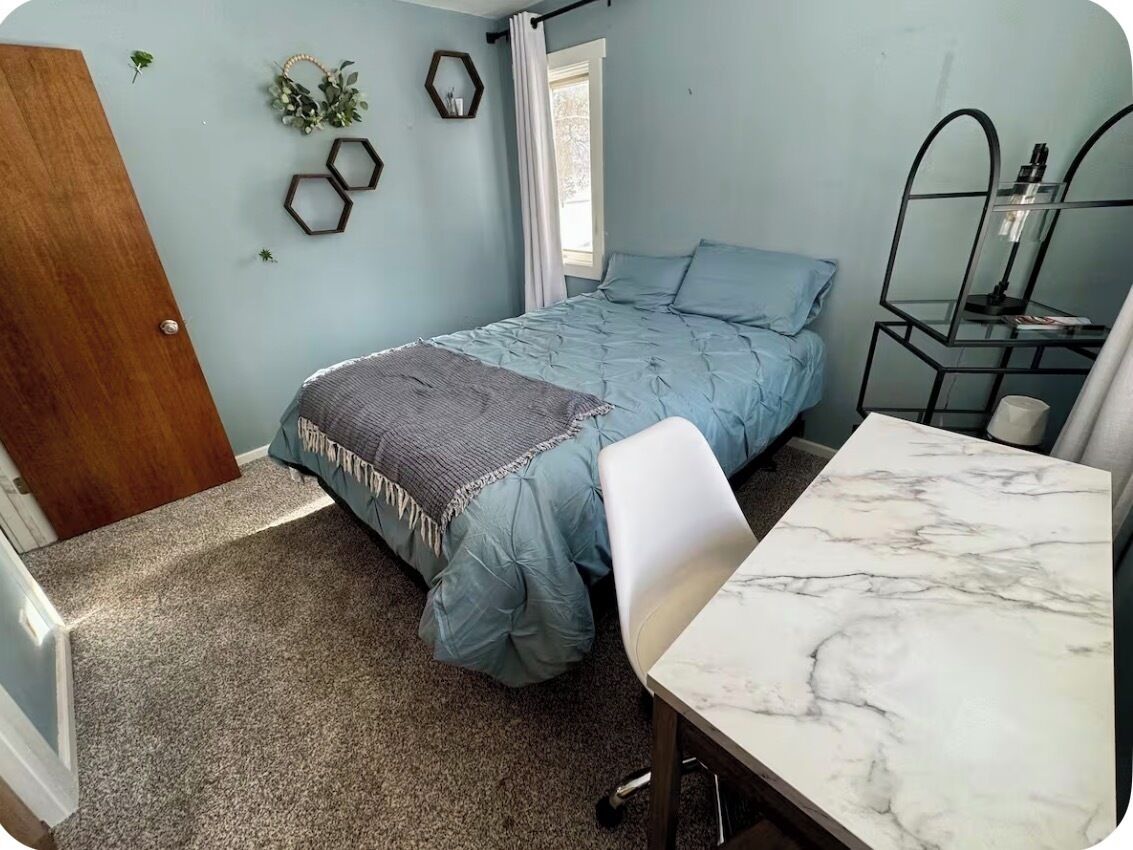 A bedroom with a bed, a desk, a chair, a window, and a door.