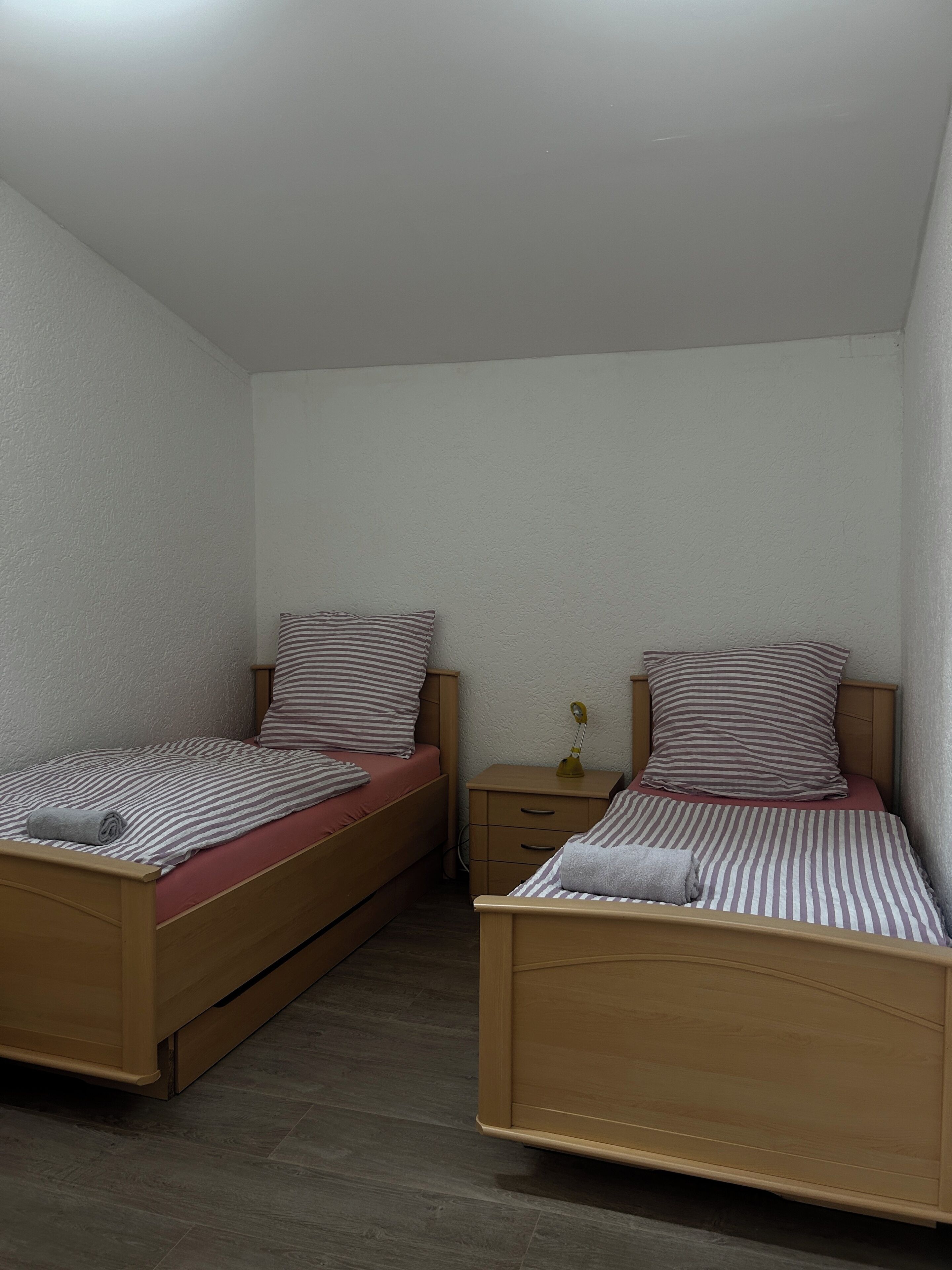 A room with two wooden beds, each with a striped pillow and a red mattress. There is a small bedside table with a lamp and a drawer.
