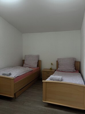 A room with two wooden beds, each with a striped pillow and a red mattress. There is a small bedside table with a lamp and a drawer. - Modern apartment that sleeps 6 (Weißenfels)
