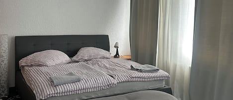 A modern bedroom with a large bed, a bedside table with a lamp, and a window with curtains.