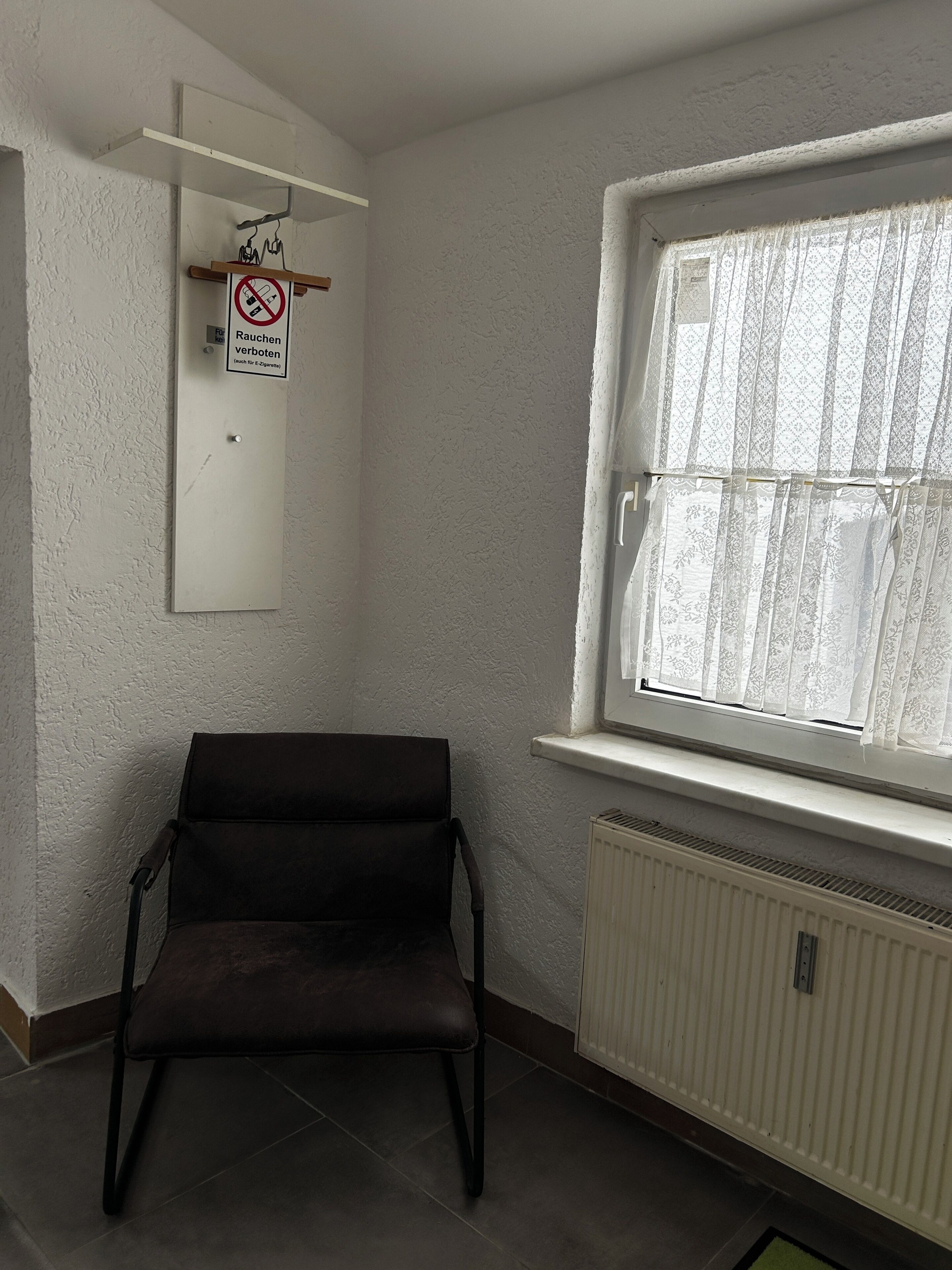 Quarto com radiador, janela com cortinas de renda, uma cadeira e um aviso proibindo o fumo.