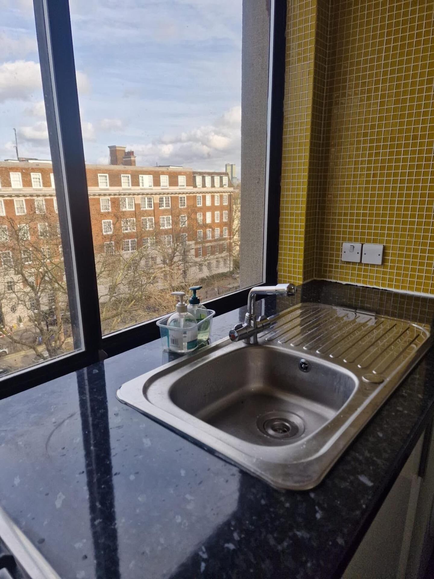 A kitchen with a stainless steel sink, a window with a view of buildings, and a yellow tiled backsplash.