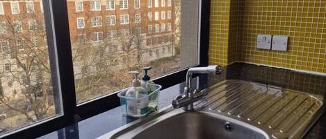 A kitchen with a stainless steel sink, a window with a view of buildings, and a yellow tiled backsplash.