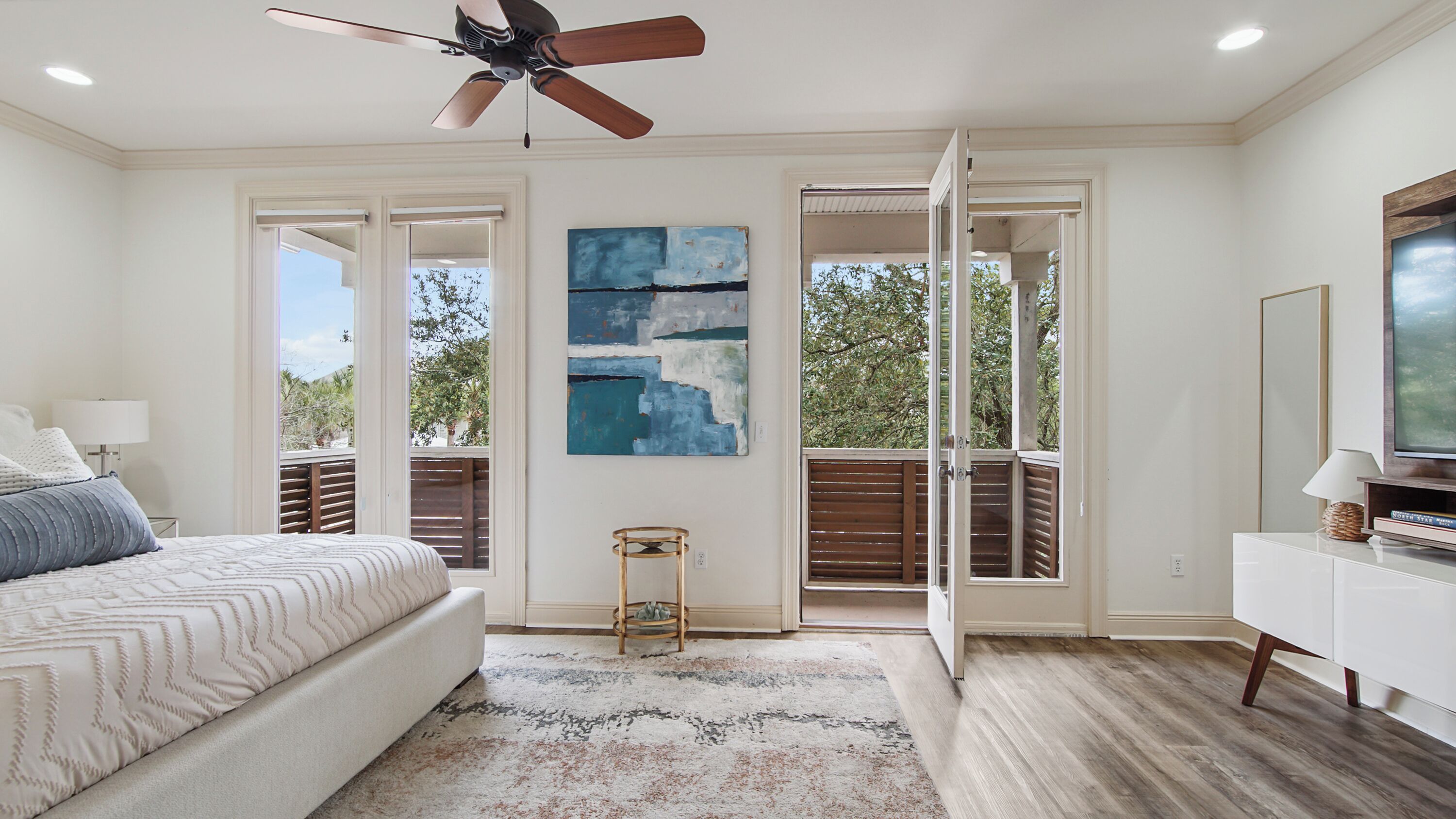 A bedroom with a bed, a ceiling fan, a painting, a window, and a view of trees.