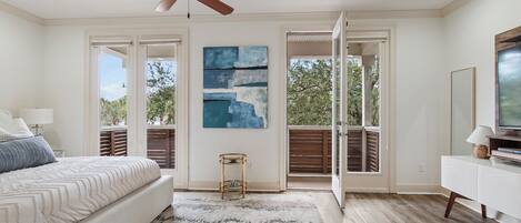 A bedroom with a bed, a ceiling fan, a painting, a window, and a view of trees.