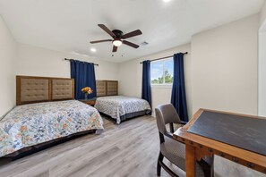 A room with two beds, a ceiling fan, a window with curtains, and a desk with a chair.