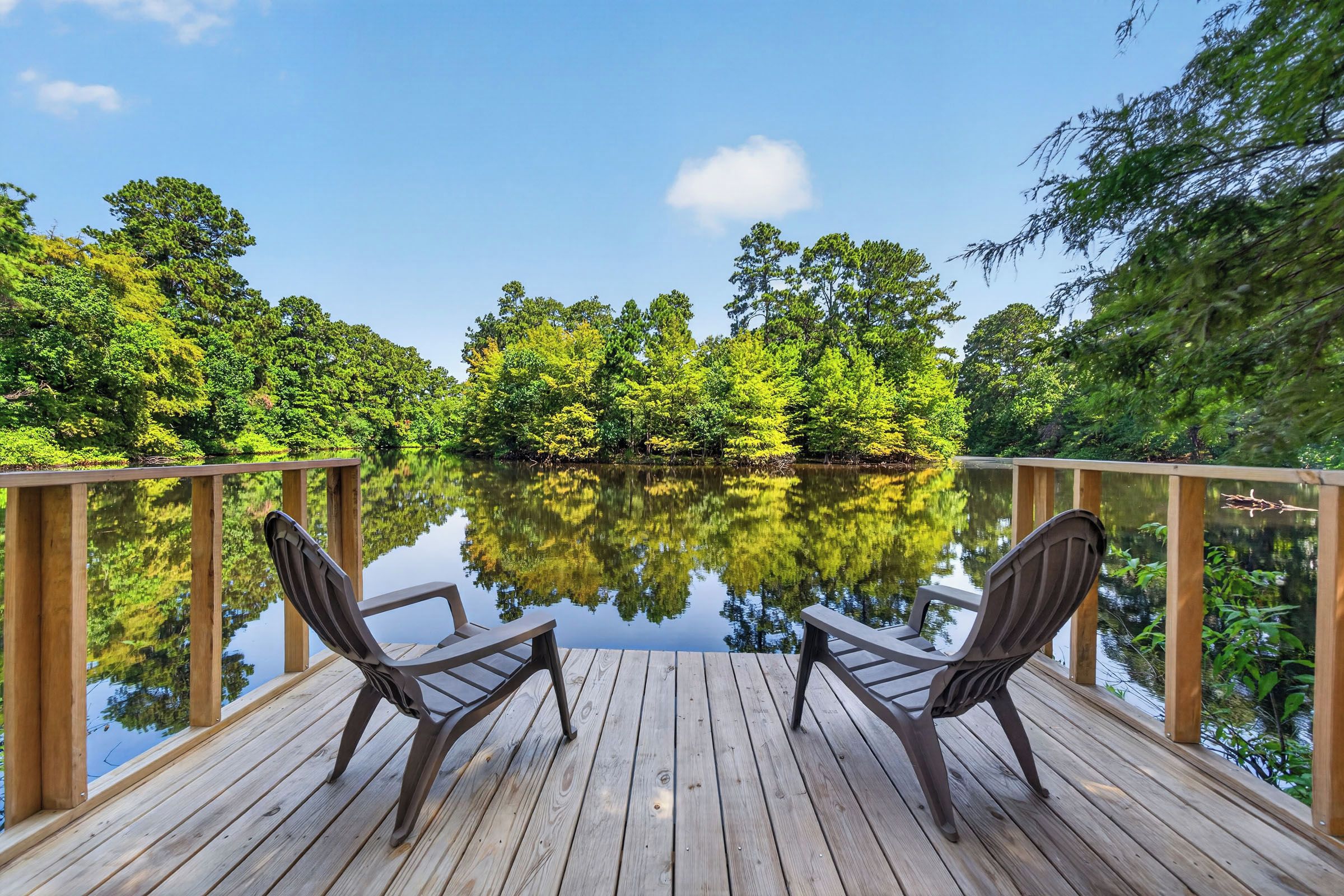 A wooden deck with two chairs facing a calm lake surrounded by dense greenery.
