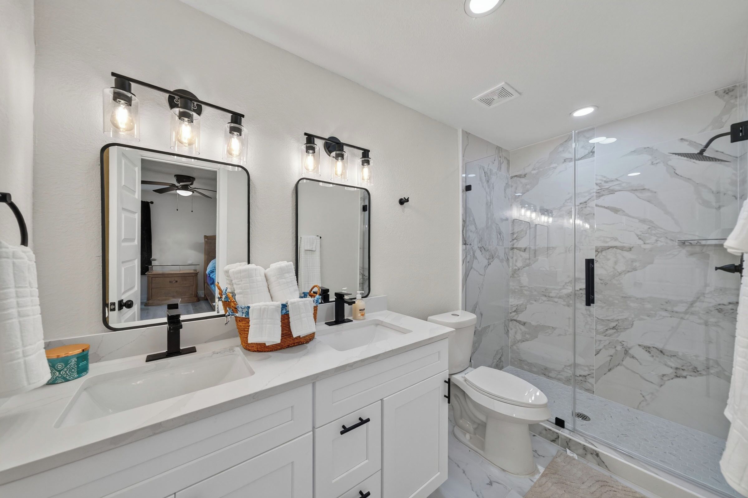 A modern bathroom with a white vanity, two mirrors, a glass shower enclosure, and marble walls.