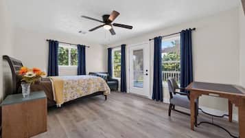 A bedroom with a bed, a desk, a chair, a ceiling fan, and a view of the outdoors through a door.
